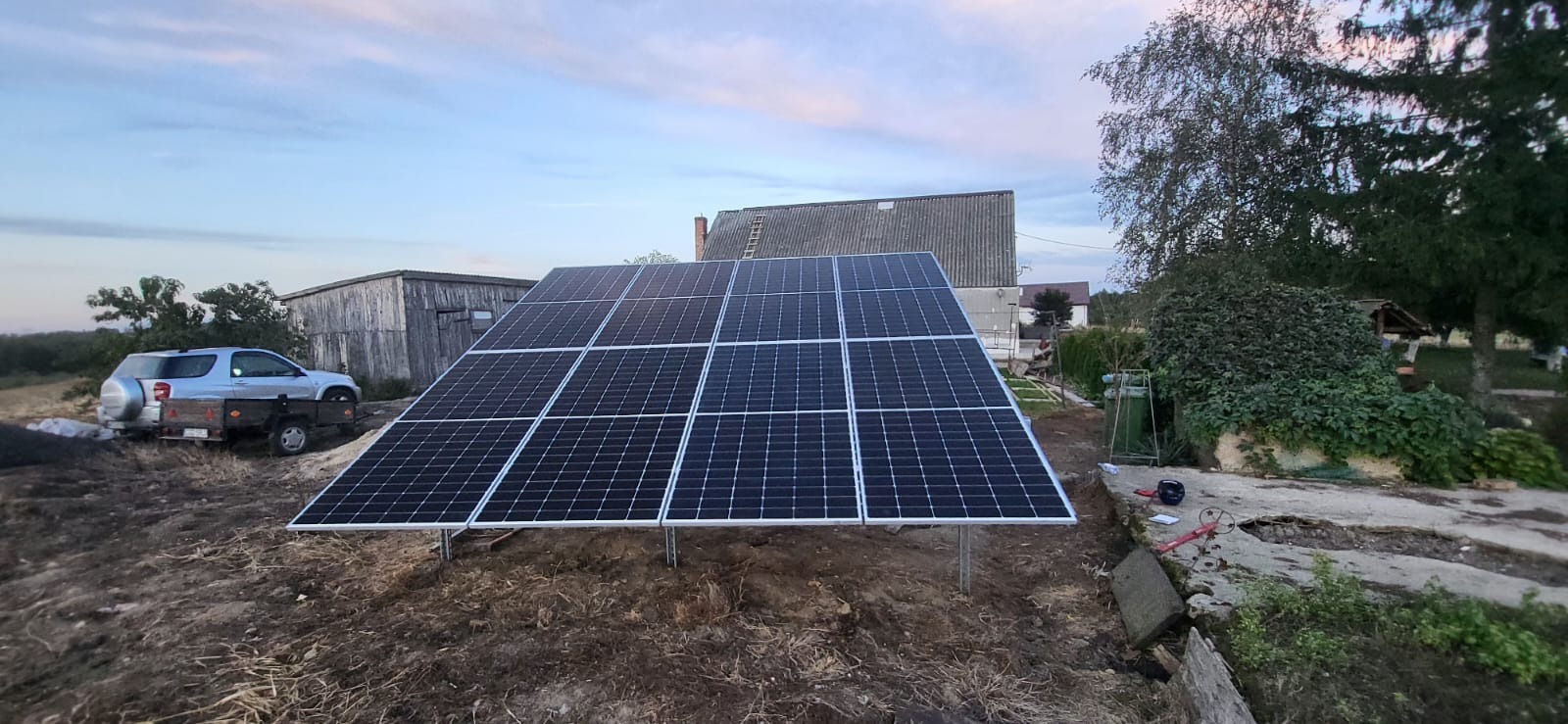 Panel słoneczny na metalowych nogach, zamontowany na nierównym terenie z widocznym samochodem z przyczepką w tle, przy zachodzącym słońcu.