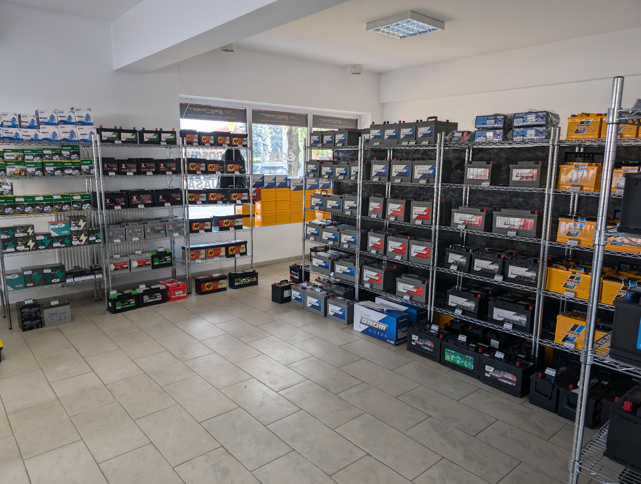 Wnętrze sklepu z akumulatorami samochodowymi, regały wypełnione różnymi modelami i markami, widok na okno i fragment ulicy.