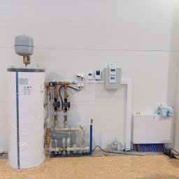 RG INSTALACJE Sp. z o.o. - Nowa instalacja centralnego ogrzewania z zasobnikiem ciepłej wody, pompami obiegowymi i rozdzielaczem podłogówki, jeszcze w folii ochronnej, zamontowana w pomieszczeniu o ścianach z paneli.