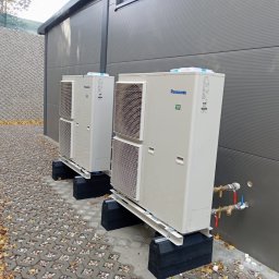 RG INSTALACJE Sp. z o.o. - Dwie zewnętrzne jednostki pompy ciepła Panasonic R32 ustawione na czarnych podstawach, z widocznymi rurami przyłączeniowymi i zaworami, na tle szarej ściany i ażurowego chodnika z opadłymi liśćmi.