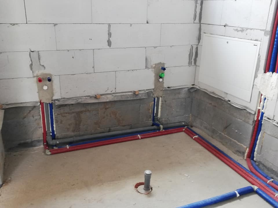Surowe wnętrze łazienki w trakcie prac hydraulicznych: widoczne przyłącza ciepłej i zimnej wody w ścianie z betonu komórkowego, rury PEX w kolorach czerwonym i niebieskim prowadzone wzdłuż podłogi...