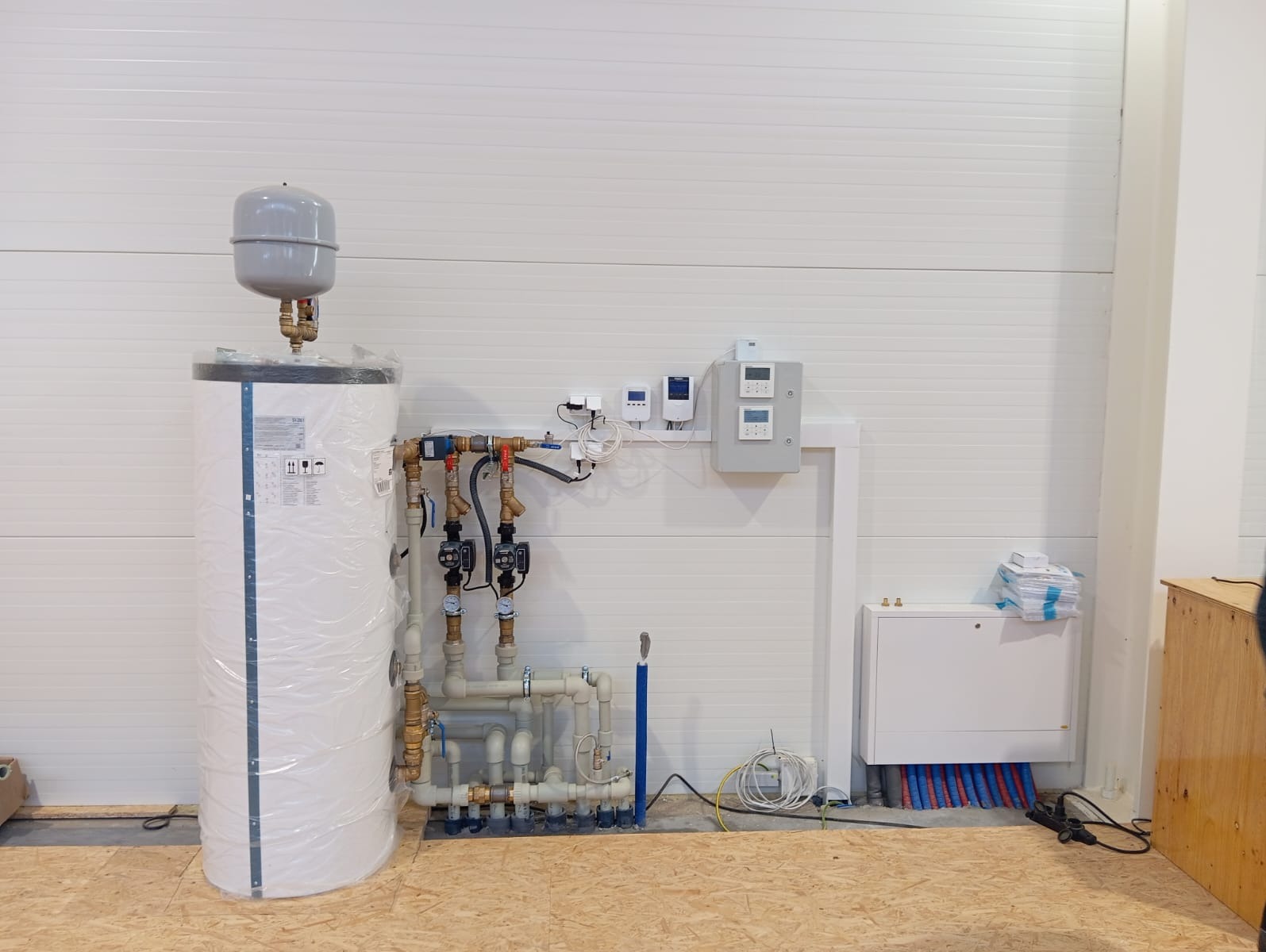 Nowa instalacja centralnego ogrzewania z zasobnikiem ciepłej wody, pompami obiegowymi i rozdzielaczem podłogówki, jeszcze w folii ochronnej, zamontowana w pomieszczeniu o ścianach z paneli.