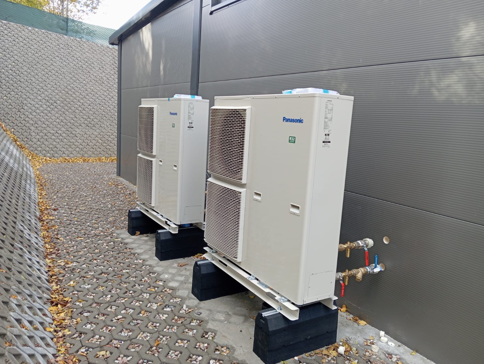 Dwie zewnętrzne jednostki pompy ciepła Panasonic R32 ustawione na czarnych podstawach, z widocznymi rurami przyłączeniowymi i zaworami, na tle szarej ściany i ażurowego chodnika z opadłymi liśćmi.