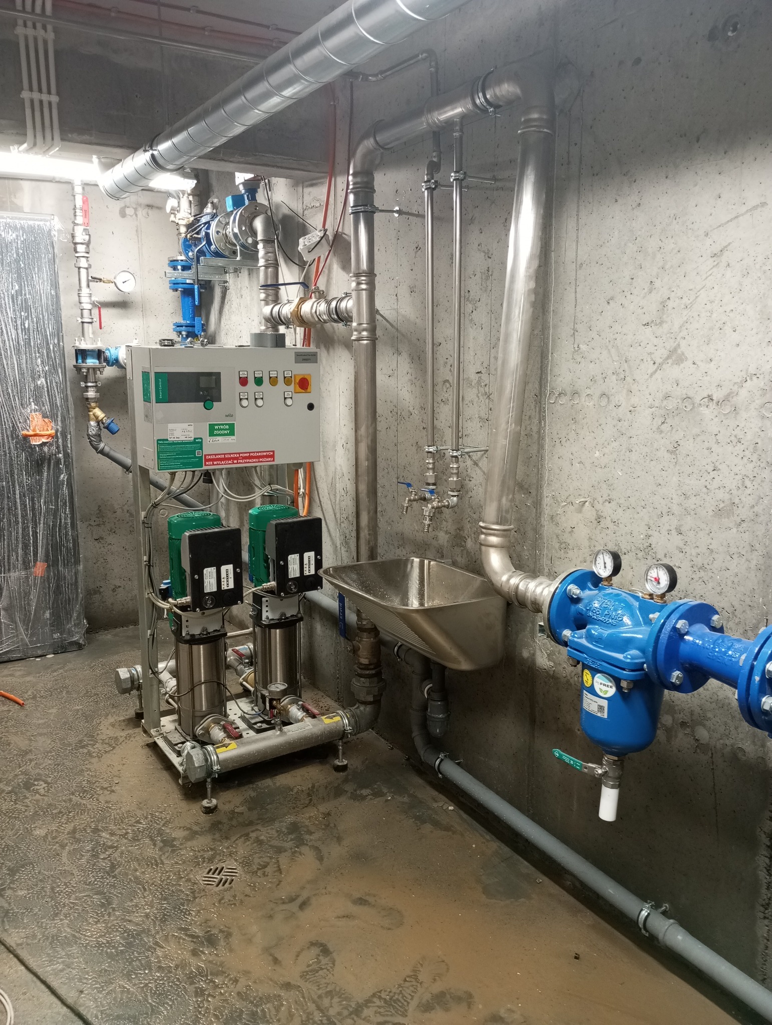 Złożona instalacja wodociągowa z dwoma pionowymi pompami, panelem sterowania z zielonymi i czerwonymi przyciskami, rurami ze stali nierdzewnej i niebieskim filtrem wody z manometrami, zamontowana...