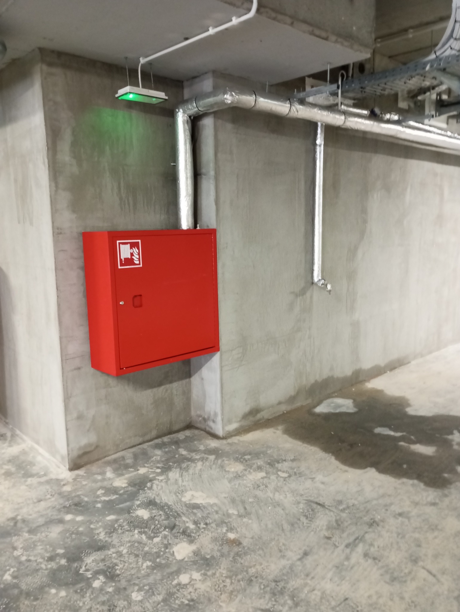 Czerwona skrzynka hydrantowa zamontowana na szarej, betonowej ścianie w pomieszczeniu przemysłowym. Nad skrzynką widoczne rury w srebrnej izolacji oraz oświetlenie awaryjne.
