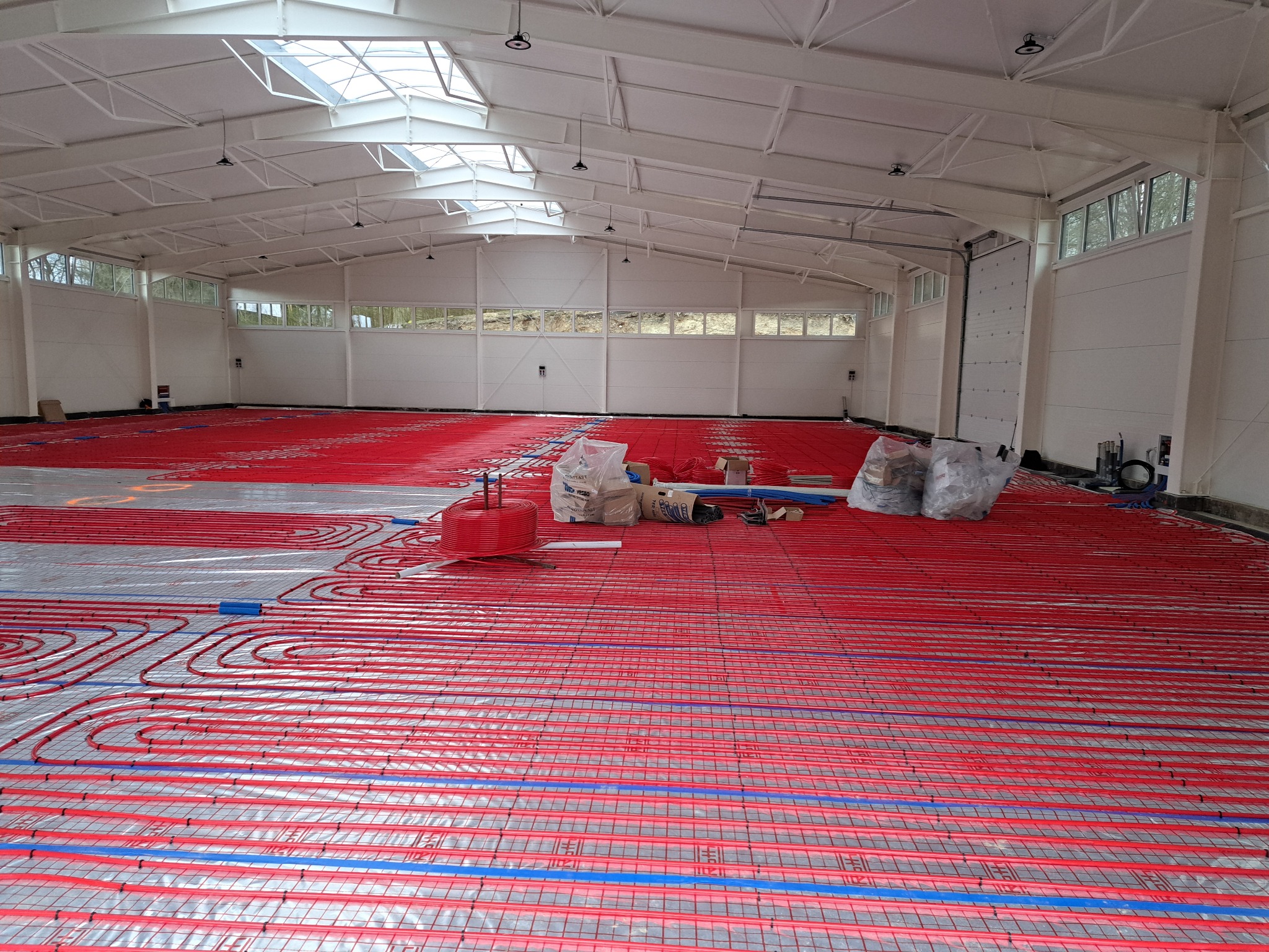 Rozległa hala z widoczną instalacją ogrzewania podłogowego: czerwone rurki ułożone w pętle na srebrnej folii, z niebieskimi elementami mocującymi. Worki z materiałami budowlanymi i narzędzia w tle,...