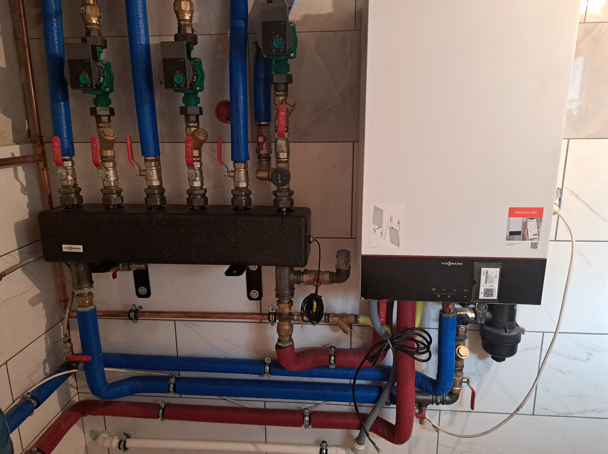 Kocioł grzewczy Viessmann z podłączonymi rurami w kolorach niebieskim i czerwonym, z widocznym rozdzielaczem i pompami obiegowymi, zamontowany na ścianie z białych płytek imitujących marmur.