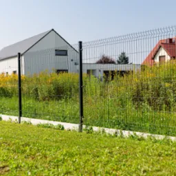 Nowoczesne ogrodzenie z siatki panelowej na betonowej podmurówce, tło z dwoma domami o różnej architekturze, bujna roślinność.
