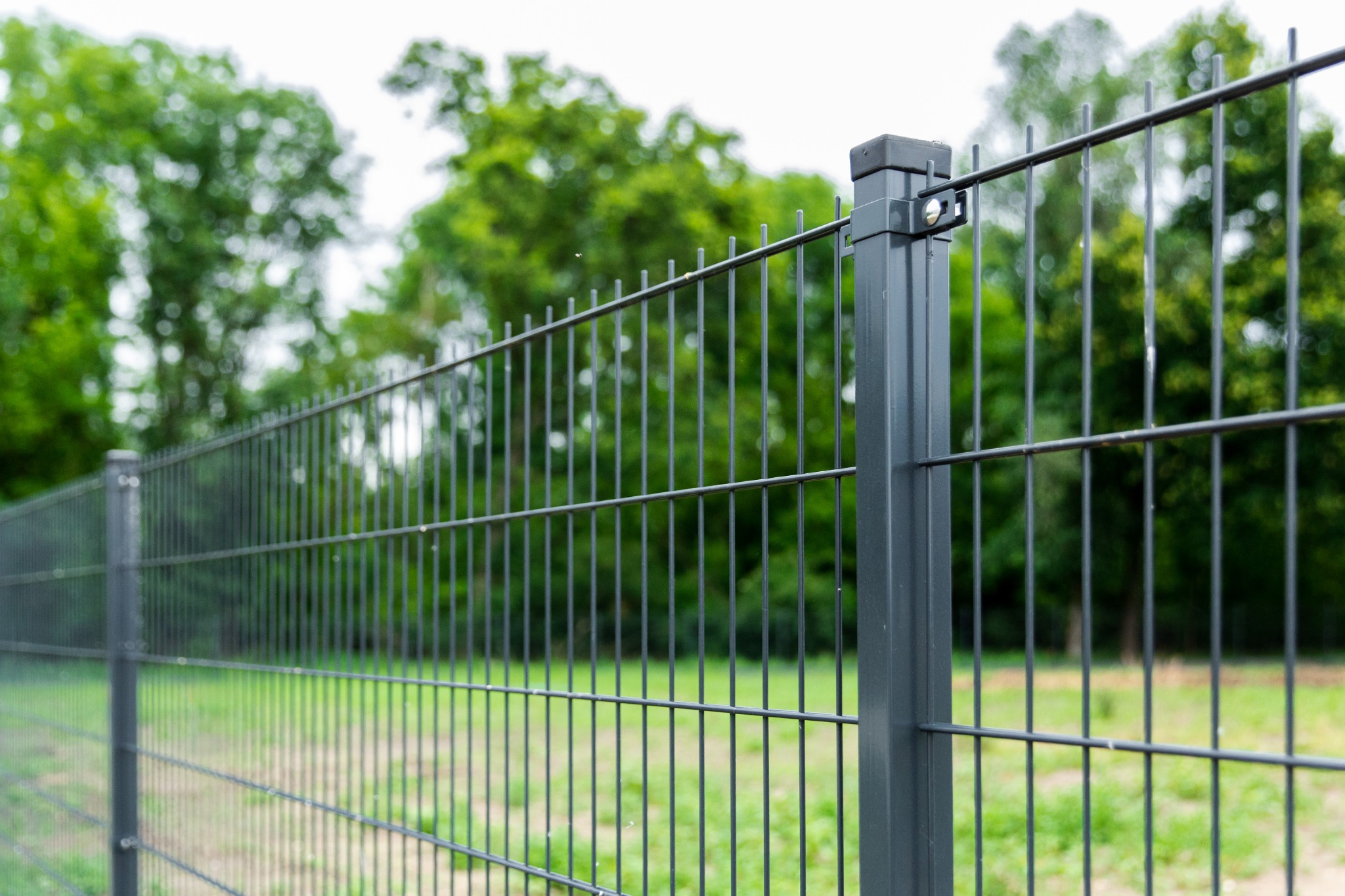 Szary, metalowy system ogrodzeniowy z paneli w Iwinach, widok z bliska na słupki i przęsła na tle zieleni drzew.