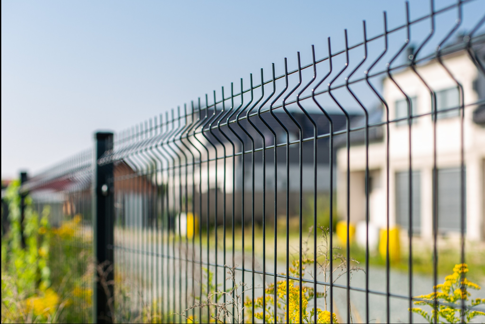 Ciemny, metalowy płot panelowy z widocznymi spawami i wystającymi drutami, na tle jasnego, dwukondygnacyjnego domu z ciemnym dachem i żółtymi akcentami, z roślinnością u podstawy.