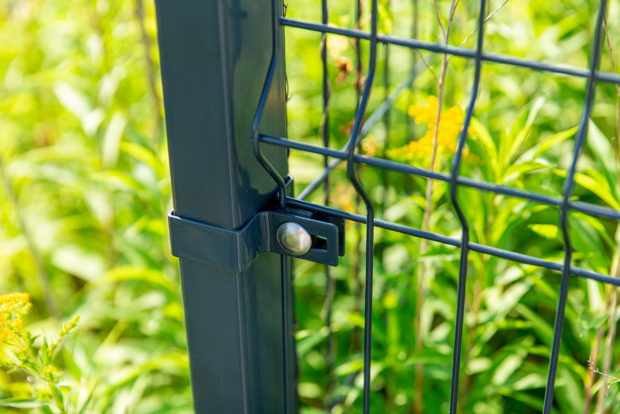 Fragment grafitowego ogrodzenia panelowego z widocznym mocowaniem słupka na tle zieleni i żółtych kwiatów.