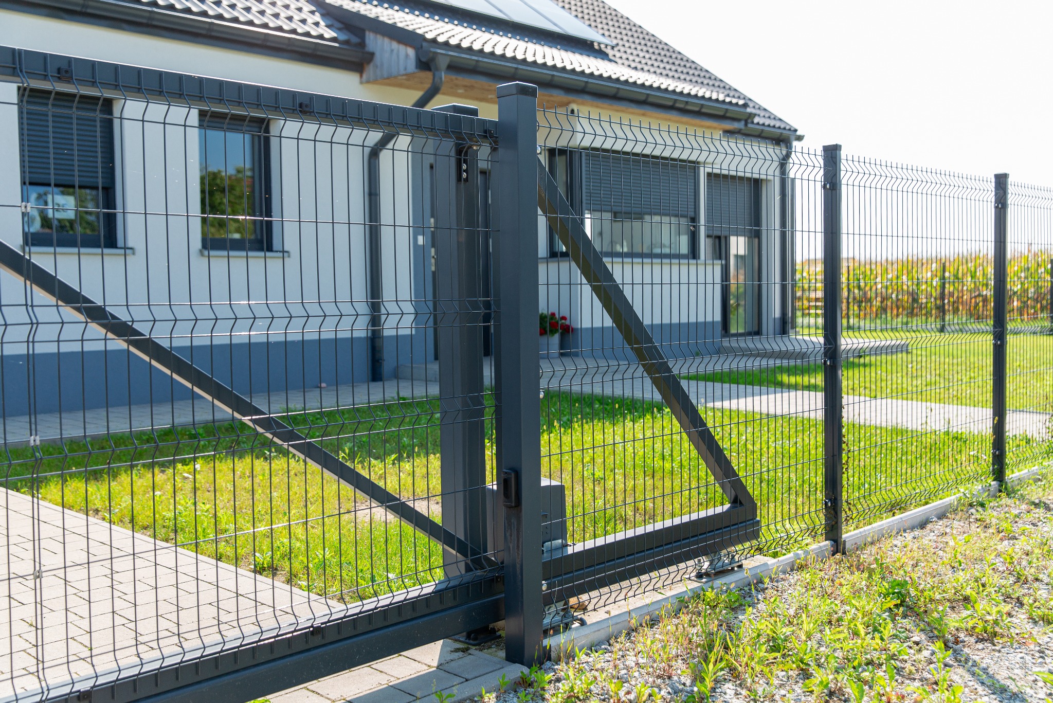 Nowoczesna posesja z przesuwną bramą wjazdową i systemem ogrodzenia panelowego w kolorze grafitowym, widok od strony zewnętrznej na tle domu z panelami słonecznymi.