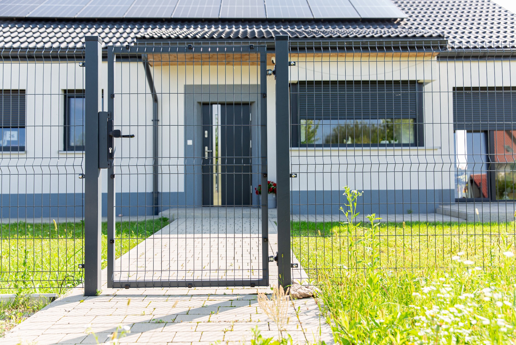 Nowoczesny dom z szarym ogrodzeniem z siatki panelowej i furtką, widok z zewnątrz na ścieżkę z kostki brukowej i bujną trawę.