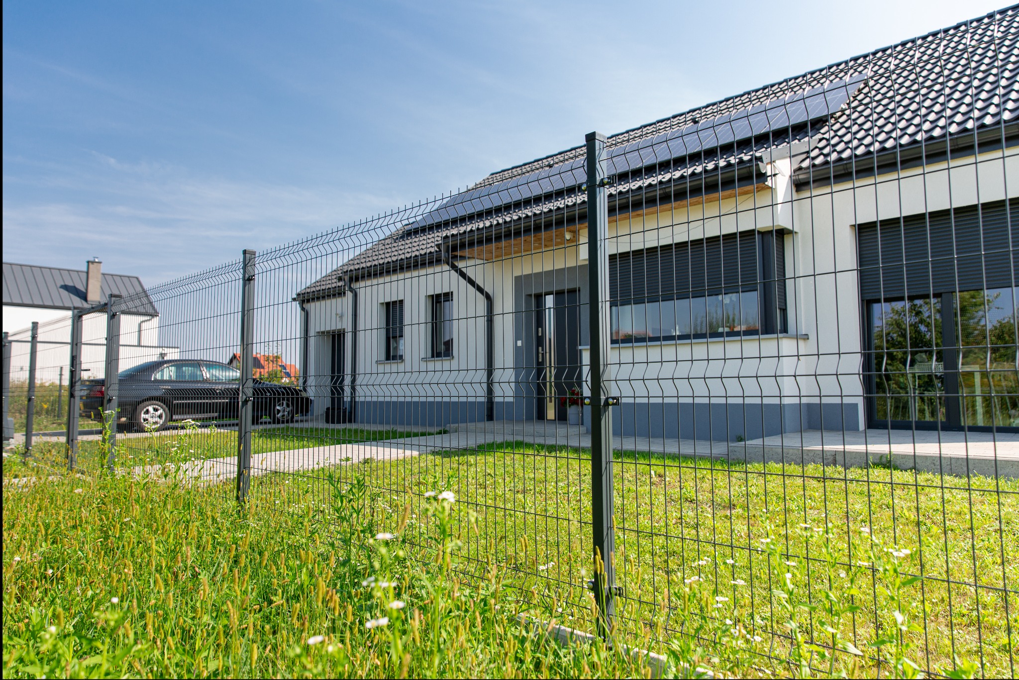 Nowoczesny dom jednorodzinny z panelami słonecznymi otoczony ciemnoszarym ogrodzeniem panelowym na tle błękitnego nieba, z widocznym samochodem osobowym za ogrodzeniem.