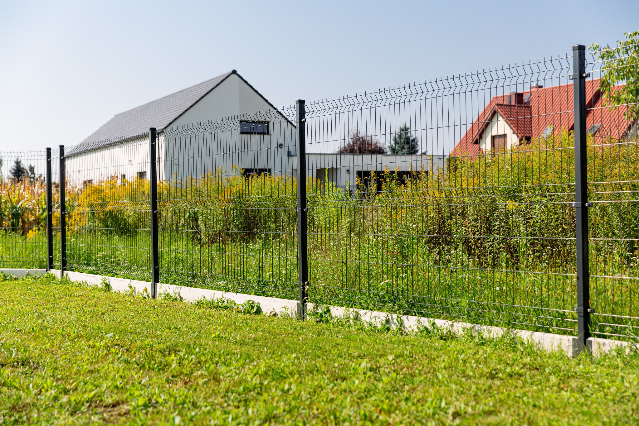 Nowoczesne ogrodzenie z siatki panelowej na betonowej podmurówce, tło z dwoma domami o różnej architekturze, bujna roślinność.