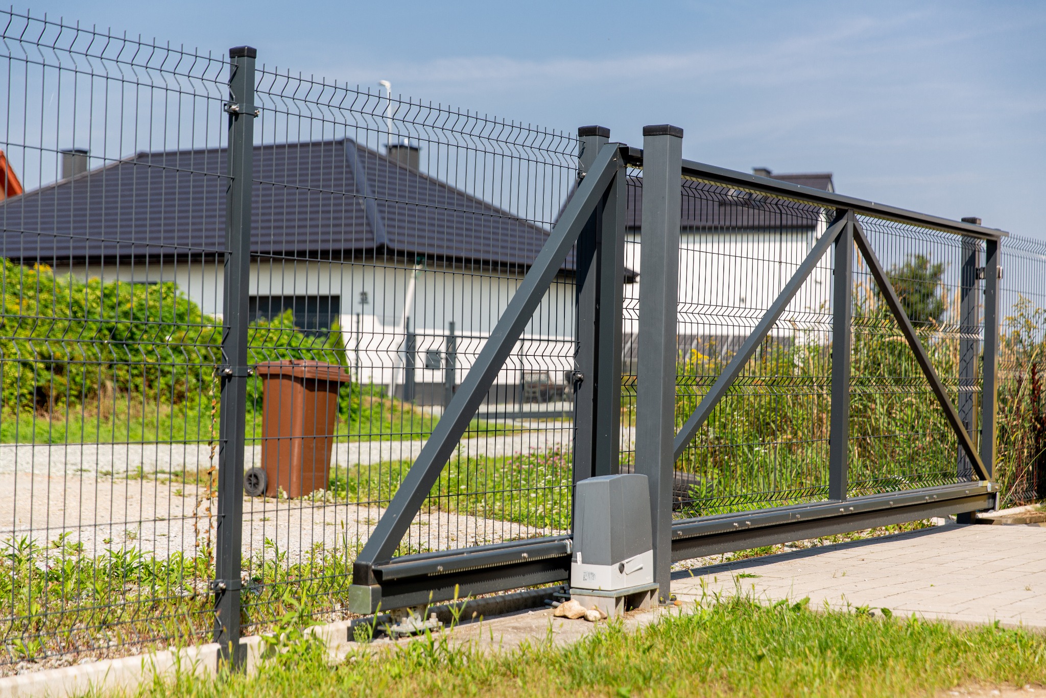Przesuwna brama z metalowej siatki z automatycznym napędem, widoczna na tle domu jednorodzinnego i fragmentu ogrodzenia panelowego.