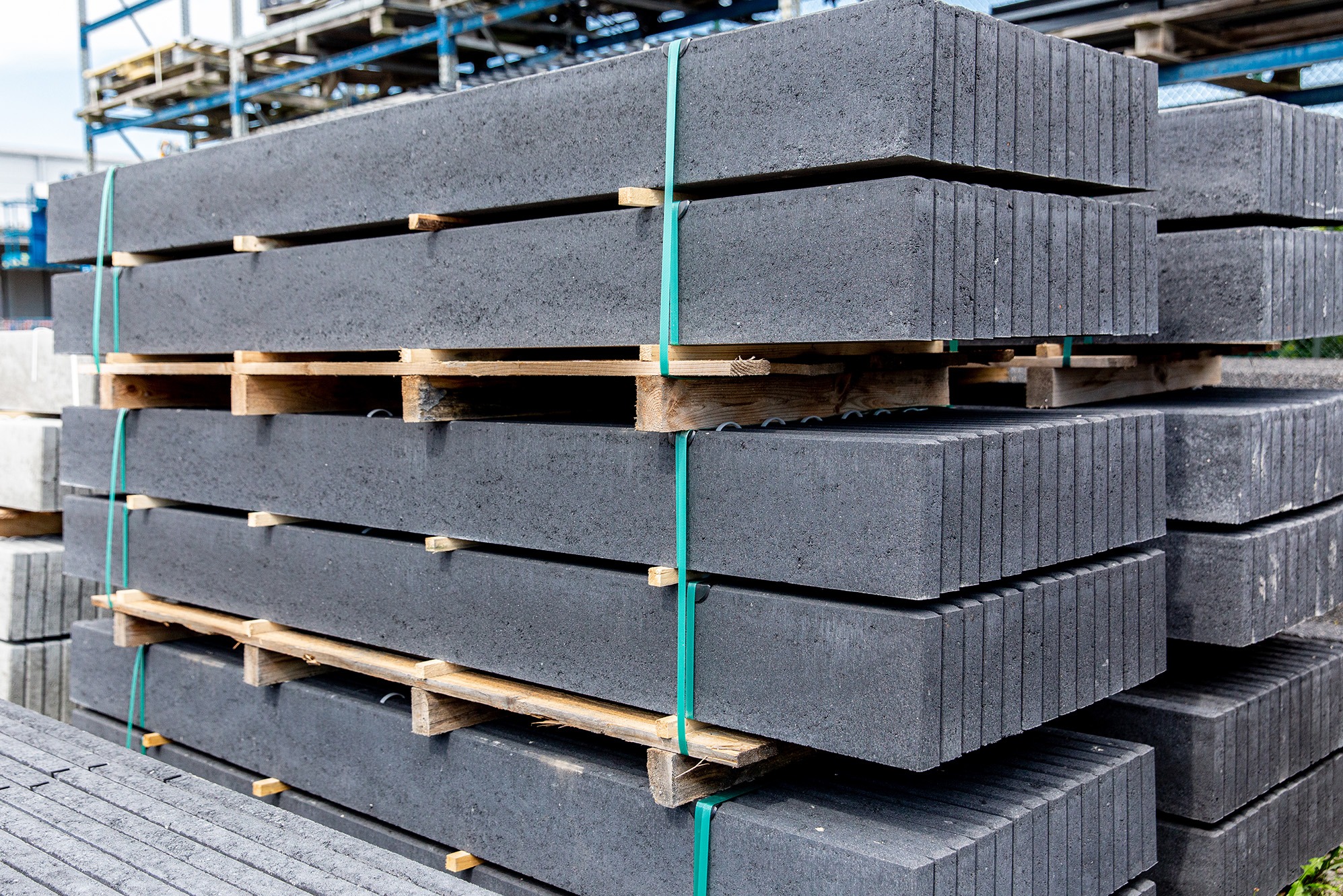 Szare betonowe elementy ogrodzeniowe ułożone na paletach, zabezpieczone zieloną taśmą, widoczne na placu składowym.