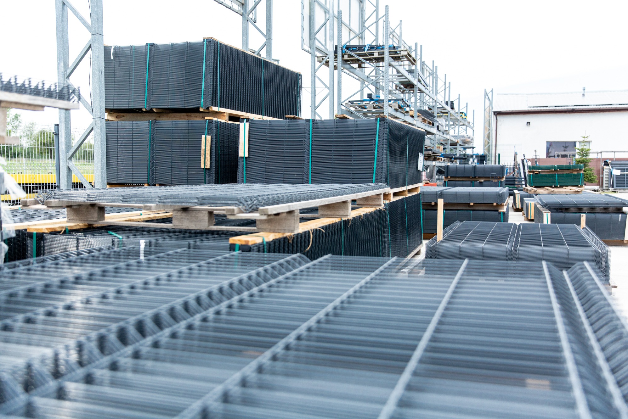 Sztaple paneli ogrodzeniowych zgrzewanych punktowo, zabezpieczonych taśmą, składowane na paletach w magazynie zewnętrznym.