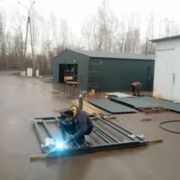 Spawanie metalowej ramy na zewnątrz, w tle budynek przemysłowy i składowane blachy, widoczny spawacz w masce i kombinezonie ochronnym.