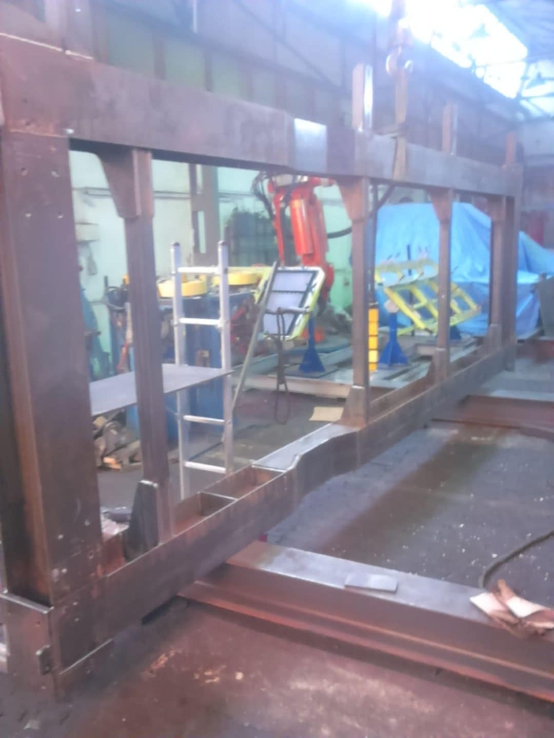 Konstrukcja stalowa w warsztacie, spawana rama z profili o przekroju prostokątnym, widoczny robot spawalniczy w tle, drabina aluminiowa i podnośnik hydrauliczny.