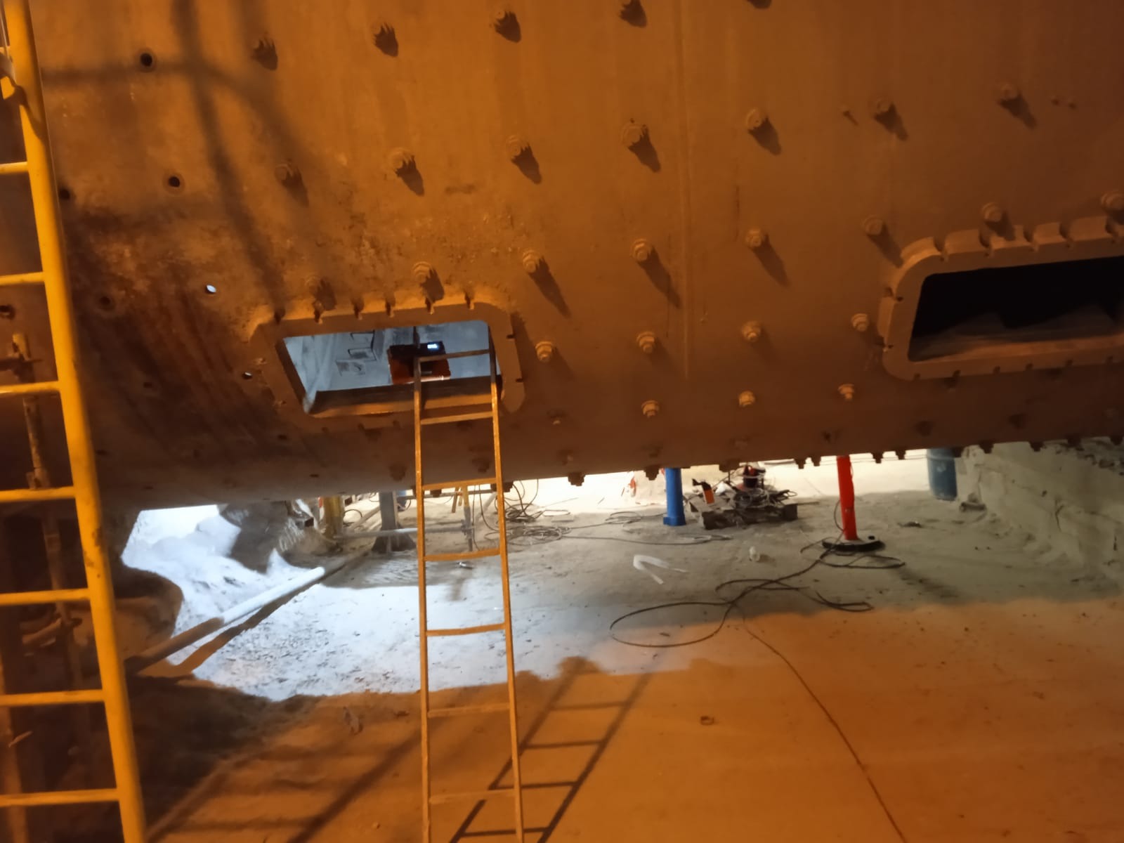 Wnętrze hali produkcyjnej z widoczną konstrukcją stalową, drabina przystawiona do otworu rewizyjnego, widoczne ślady prac spawalniczych i narzędzia.