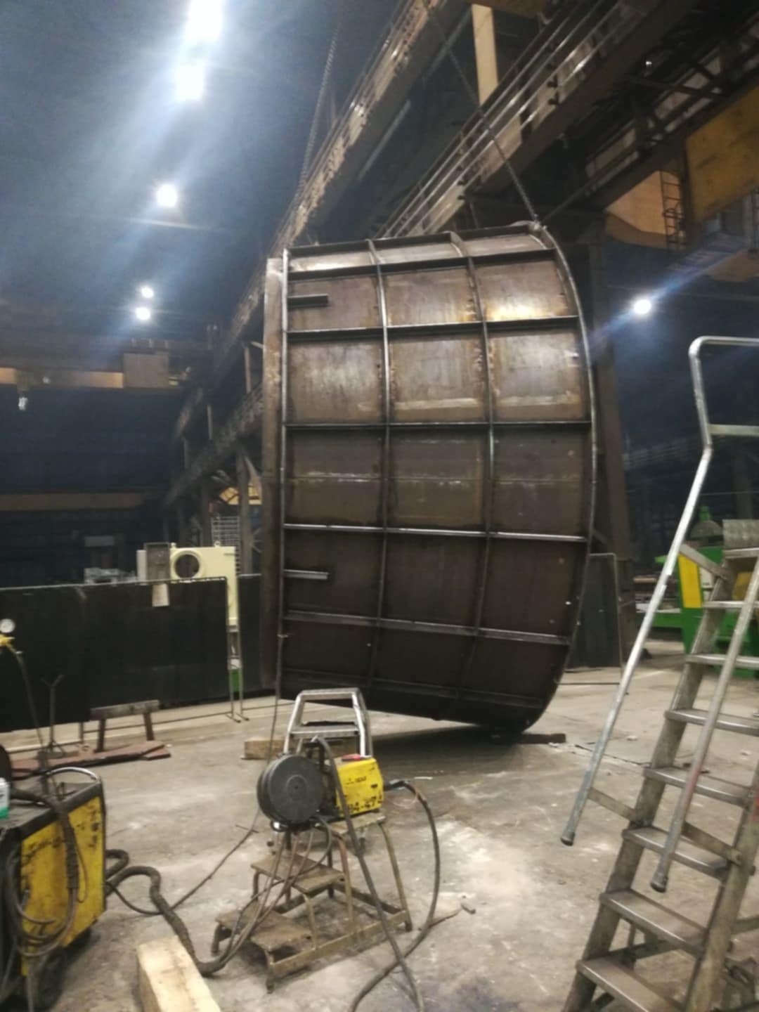 Wnętrze hali produkcyjnej z suwnicą podnoszącą dużą, metalową konstrukcję o zaokrąglonym kształcie, spawarka i drabina w pobliżu.