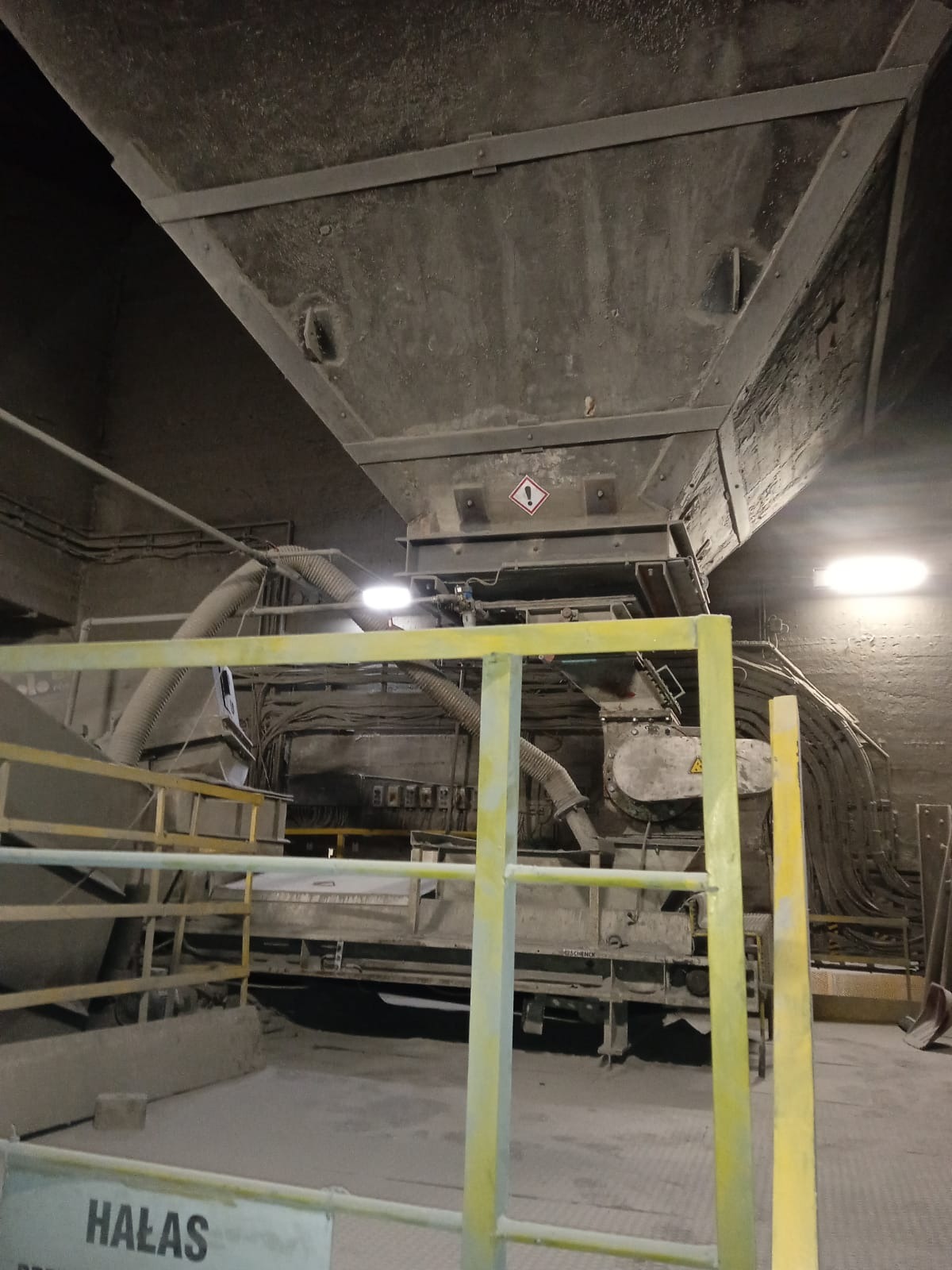 Wnętrze hali przemysłowej z metalową konstrukcją pod sufitem, rurami i żółtą barierką ochronną z napisem 'HAŁAS'.