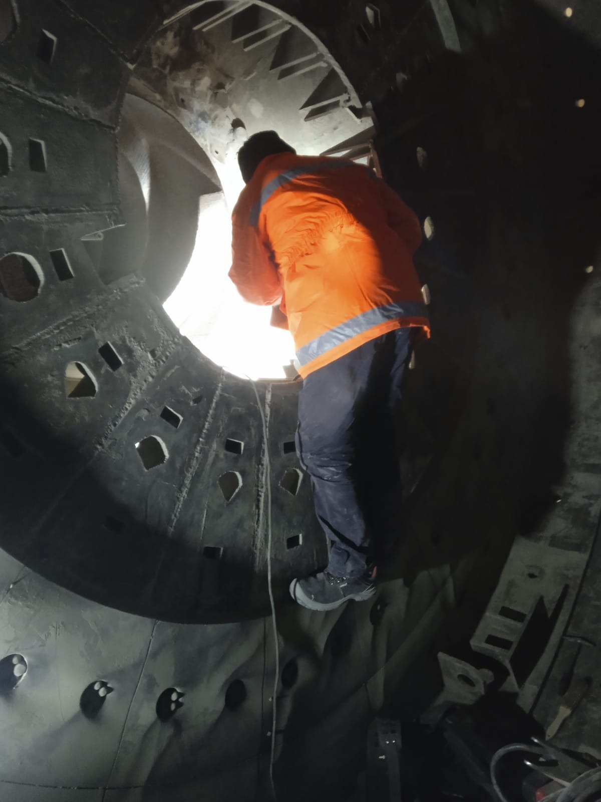 Pracownik w pomarańczowej kurtce inspekcjonuje wnętrze dużej, metalowej konstrukcji z otworami, oświetlonej od wewnątrz.