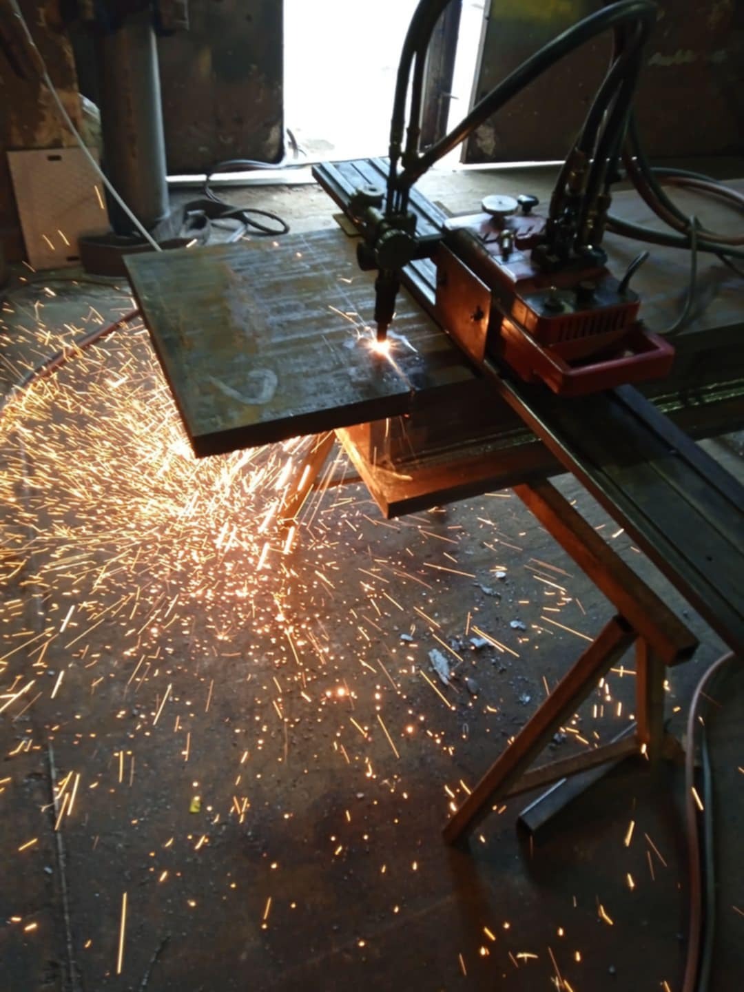 Cięcie grubego arkusza metalu za pomocą palnika acetylenowo-tlenowego, iskry rozbryzgują się na boki, widoczne elementy maszyny do cięcia.