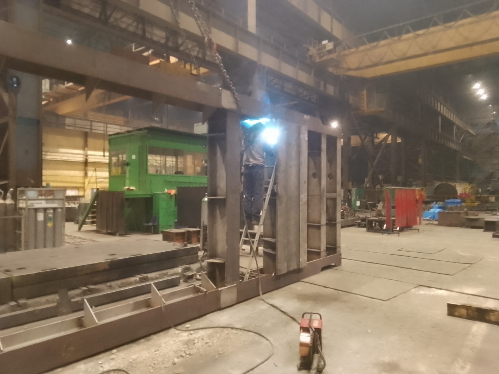 Spawanie metalowej konstrukcji szkieletowej w hali produkcyjnej, spawacz na drabinie, widoczne iskry i sprzęt spawalniczy.