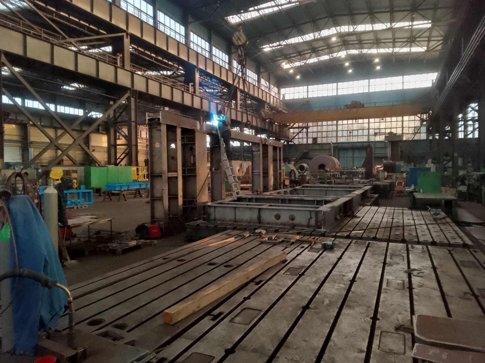 Spawanie konstrukcji stalowej na hali produkcyjnej, widoczny spawacz na drabinie, dźwig suwnicowy i elementy konstrukcyjne w trakcie montażu.