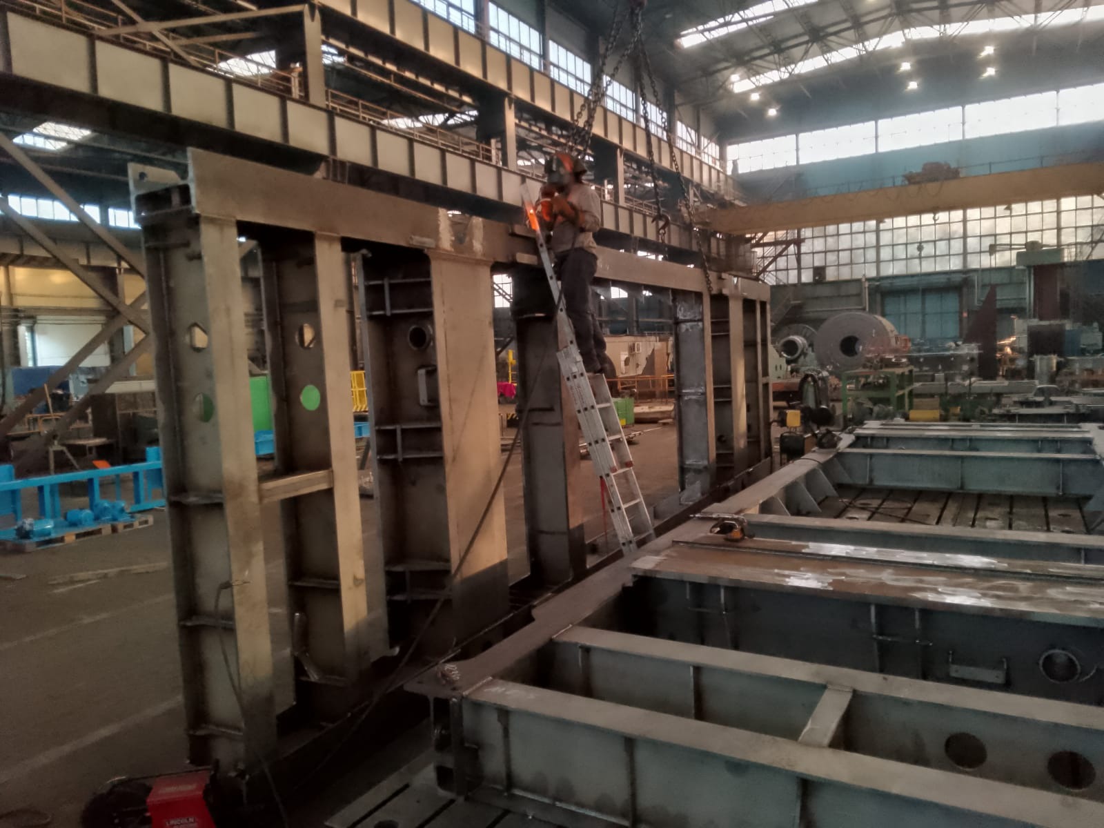 Pracownik w masce spawalniczej spawa metalową konstrukcję z drabiny w hali produkcyjnej, widoczne elementy suwnicy i inne konstrukcje stalowe w tle.