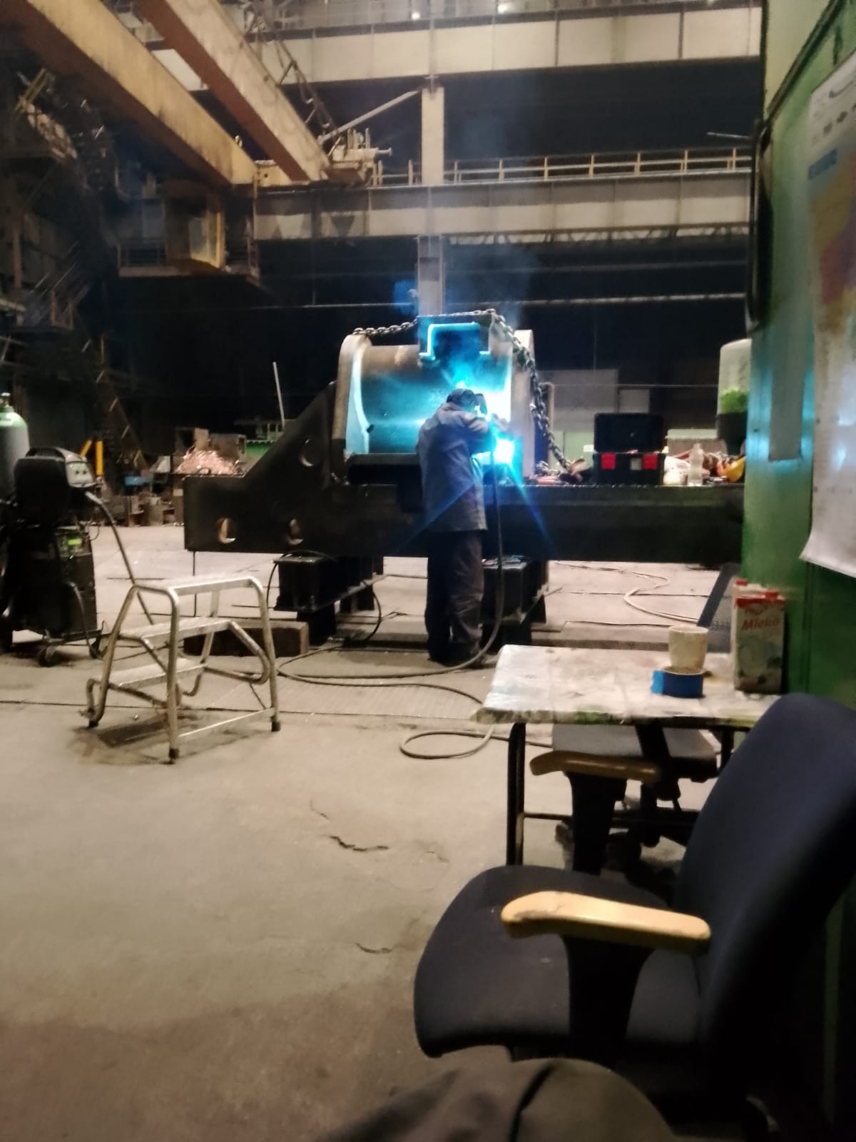 Pracownik w kombinezonie spawalniczym spawający element konstrukcji stalowej w hali produkcyjnej, widoczne iskry i narzędzia pracy, w tle suwnica i inne elementy konstrukcyjne.