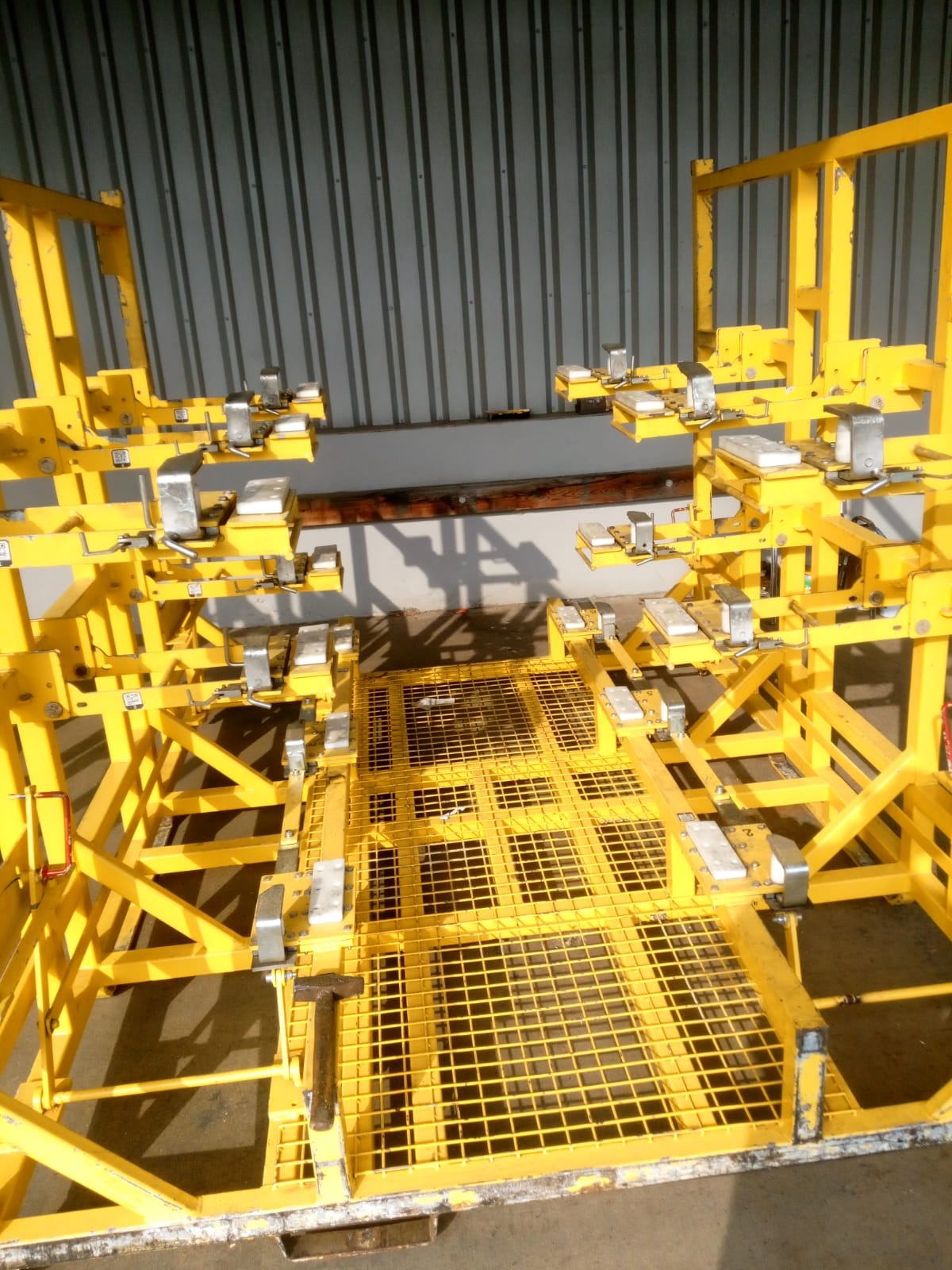 Żółta, metalowa konstrukcja szkieletowa z elementami mocującymi, prawdopodobnie używana w procesie produkcyjnym lub montażowym, widoczna młotek na siatce podłogowej, tło stanowi blaszana ściana.