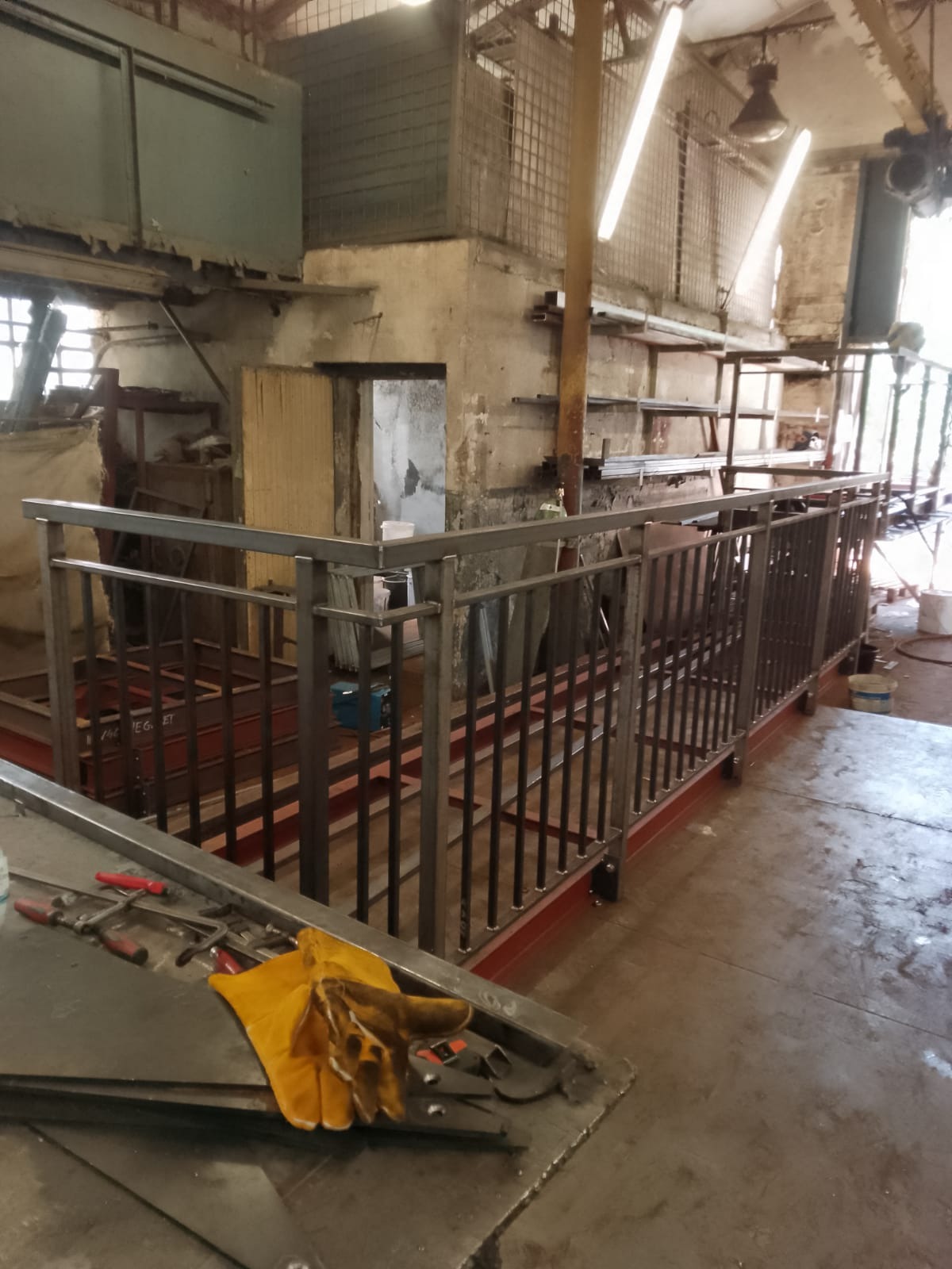 Metalowa balustrada w surowym, industrialnym wnętrzu warsztatu, z żółtymi rękawicami roboczymi i narzędziami na pierwszym planie.