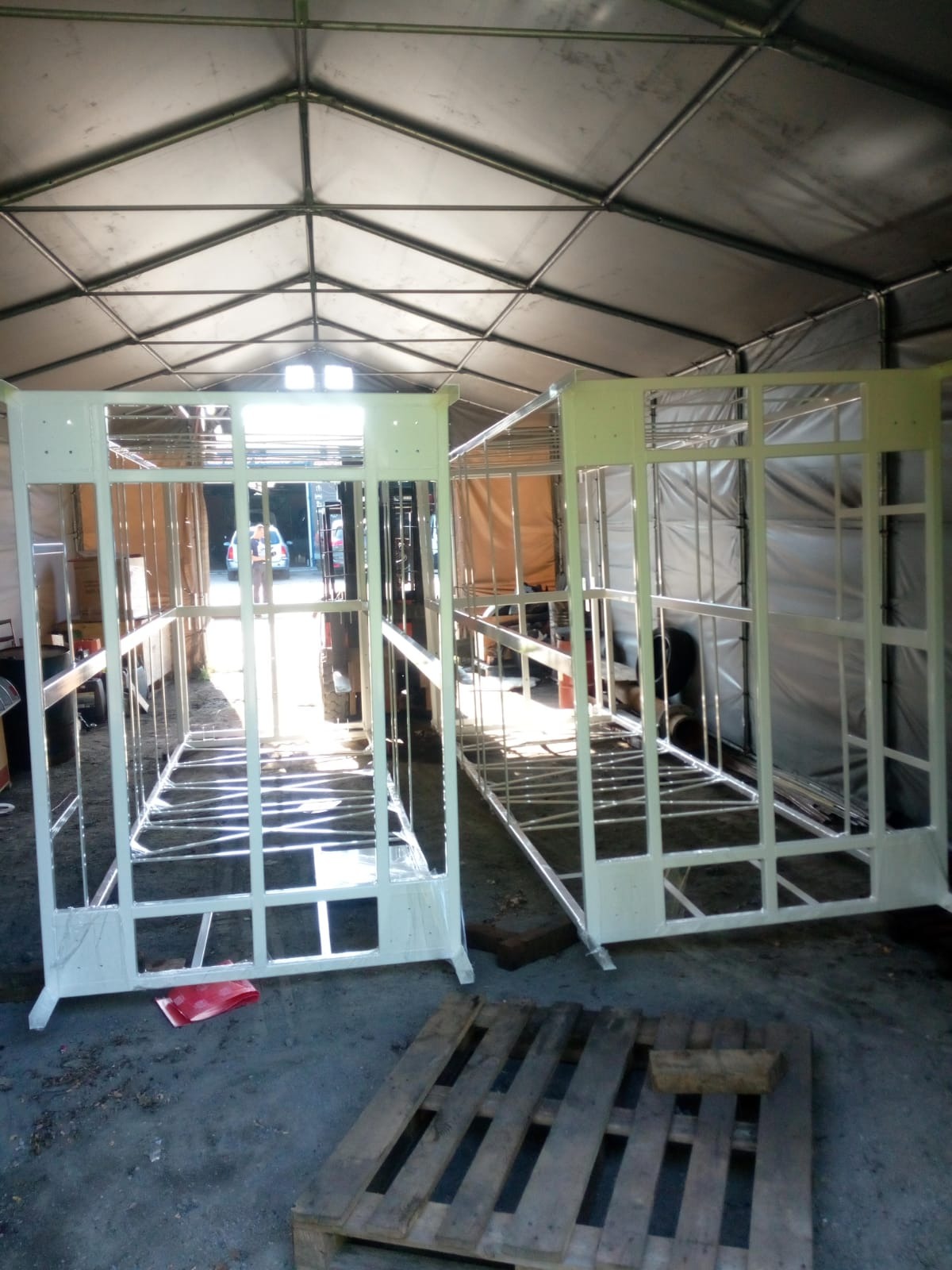 Dwie metalowe konstrukcje pomalowane na biało, prawdopodobnie fragmenty hali, w trakcie budowy wewnątrz namiotu, widoczna paleta i folia ochronna.