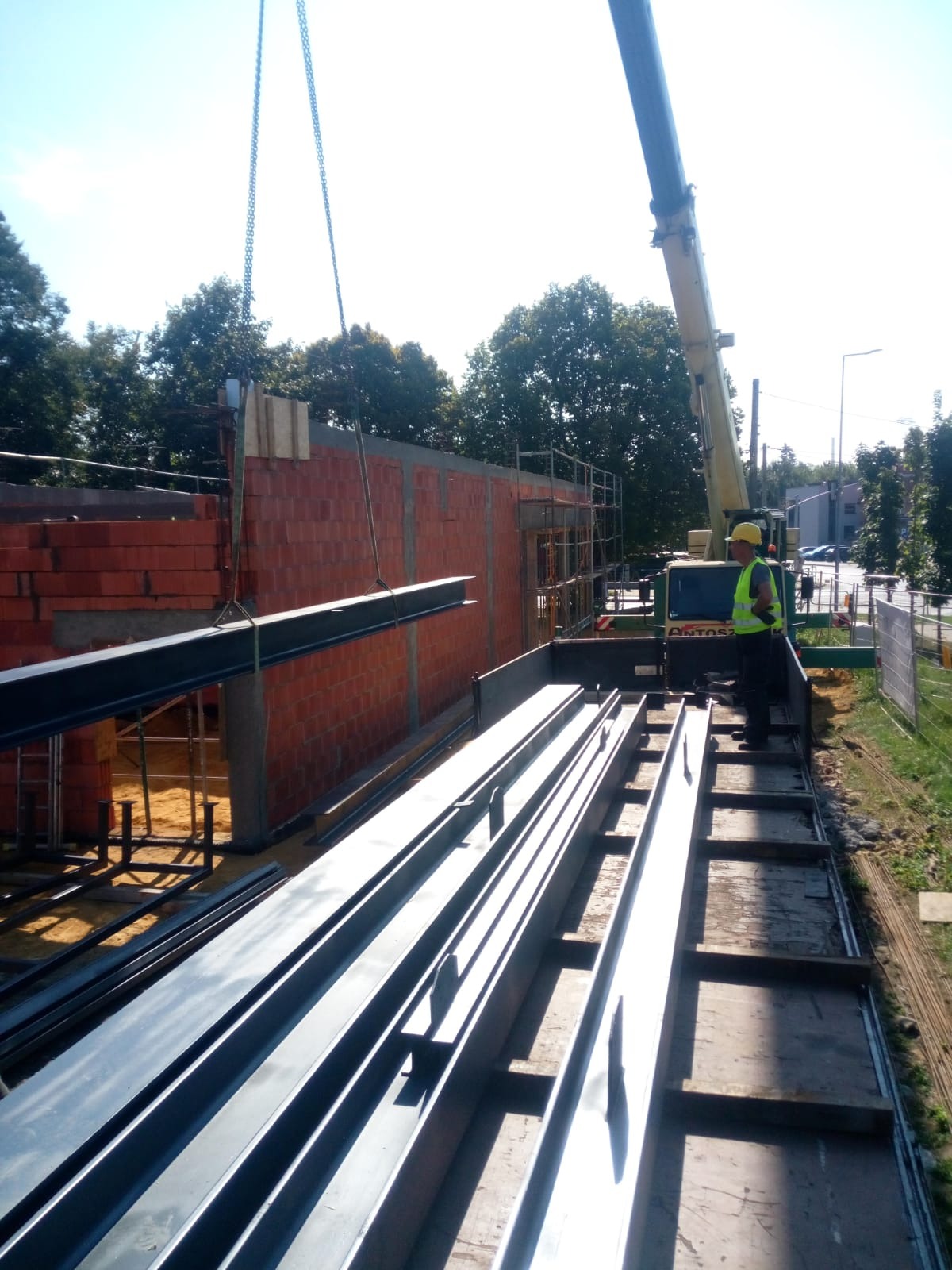 Montaż stalowej konstrukcji hali: dźwig podnosi belkę, pracownik w kasku i kamizelce odblaskowej nadzoruje proces, w tle mur z czerwonej cegły i drzewa.