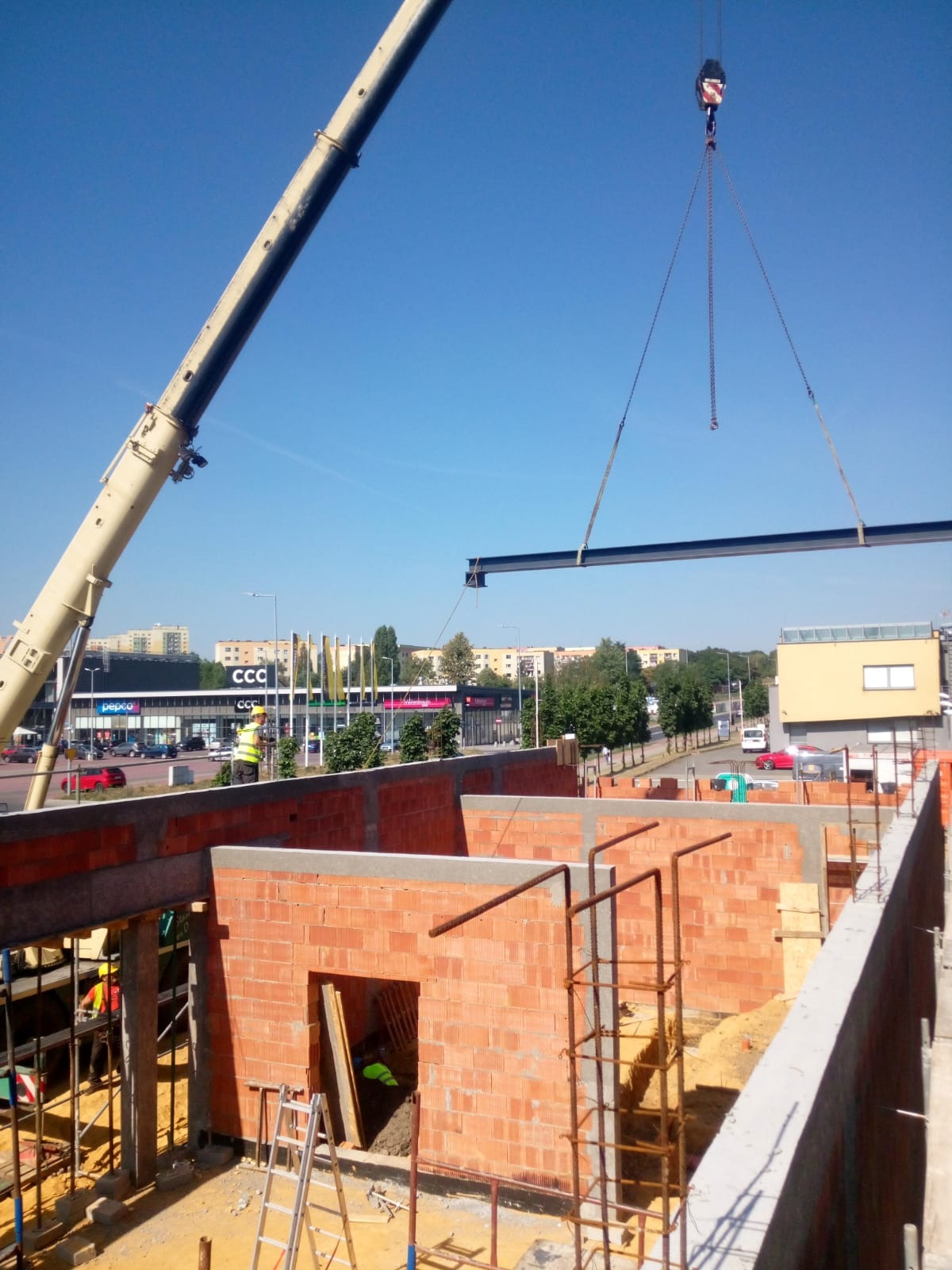 Montaż stalowej belki konstrukcyjnej na budowie budynku z cegły, widoczny dźwig i pracownicy w odblaskowych kamizelkach, w tle zabudowa miejska.