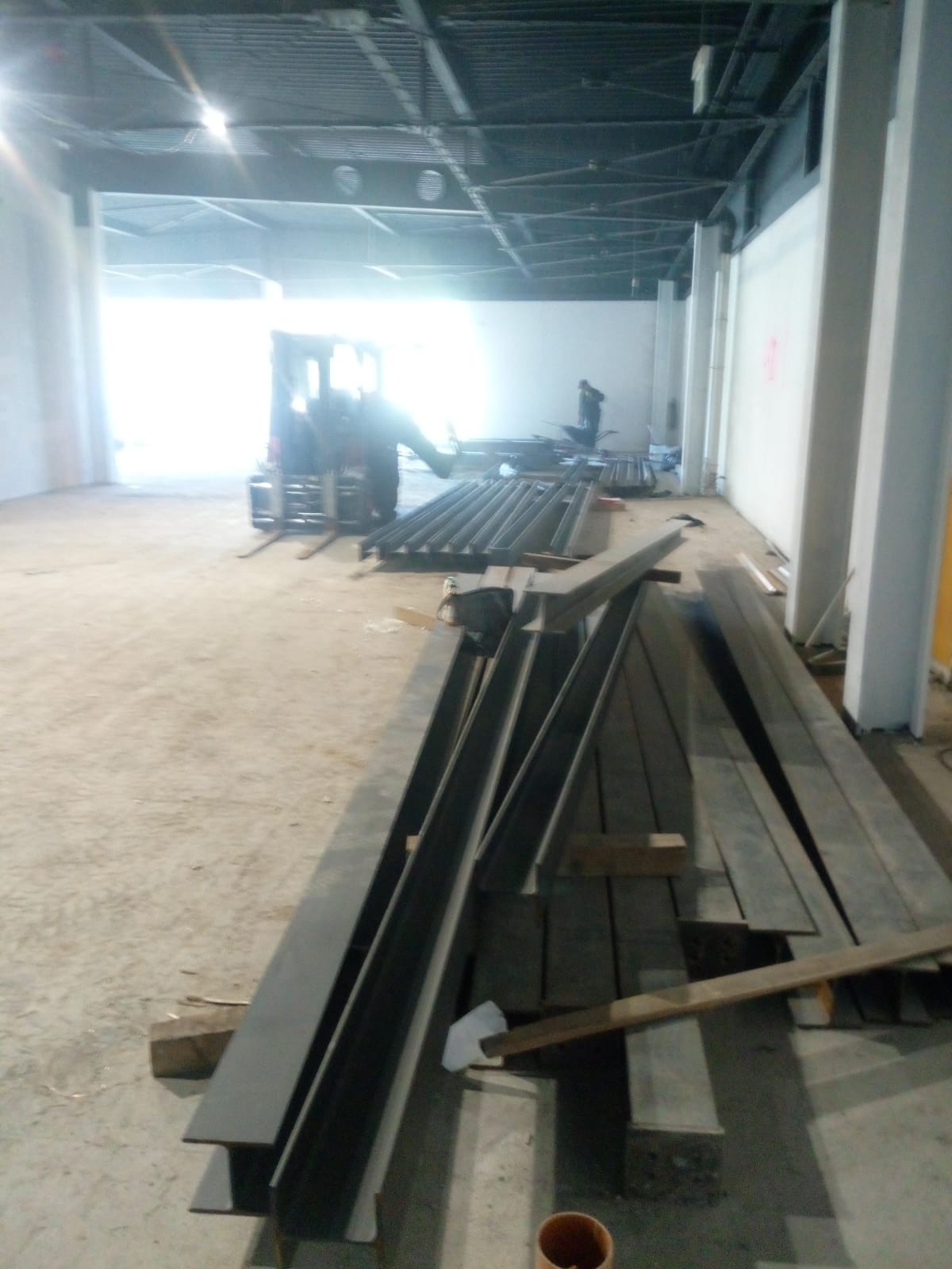 Stalowe profile konstrukcyjne ułożone na podłodze w hali w trakcie budowy, w tle wózek widłowy i pracownik.