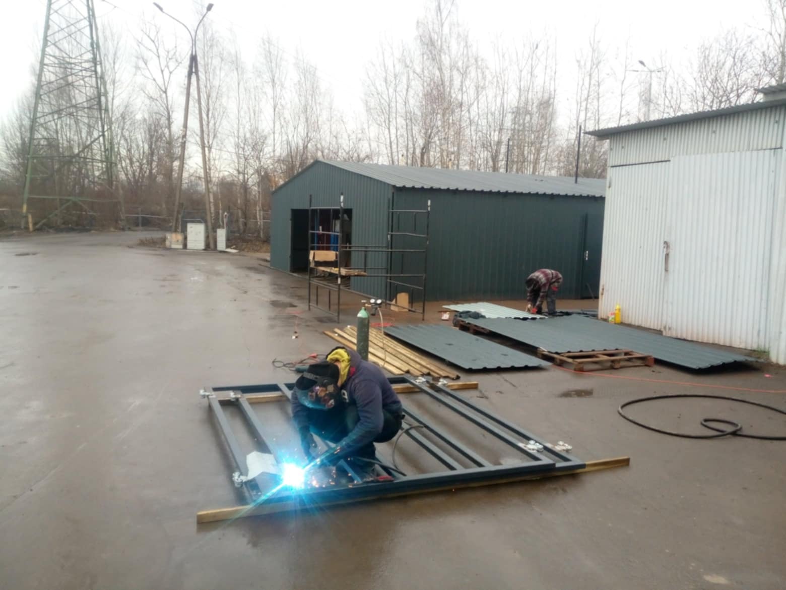 Spawanie metalowej ramy na zewnątrz, w tle budynek przemysłowy i składowane blachy, widoczny spawacz w masce i kombinezonie ochronnym.