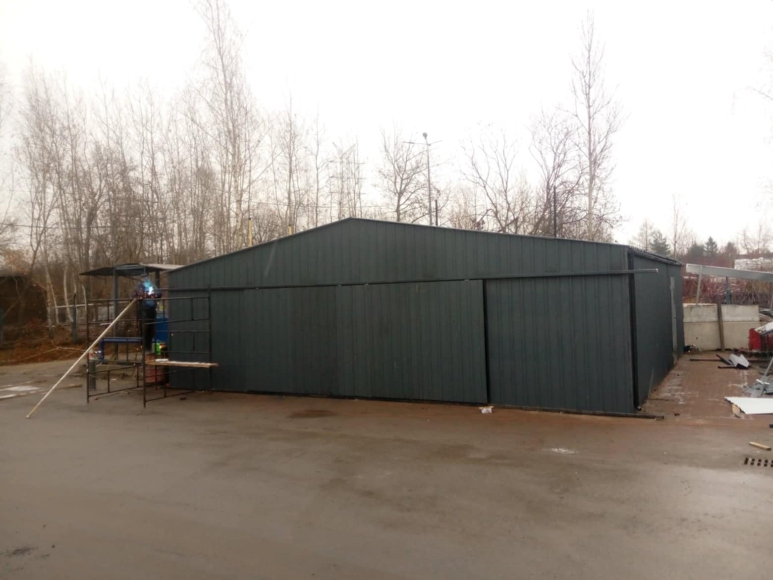 Montaż szarej hali z blachy trapezowej, spawanie konstrukcji na zewnątrz, widoczne rusztowanie i drzewa w tle.