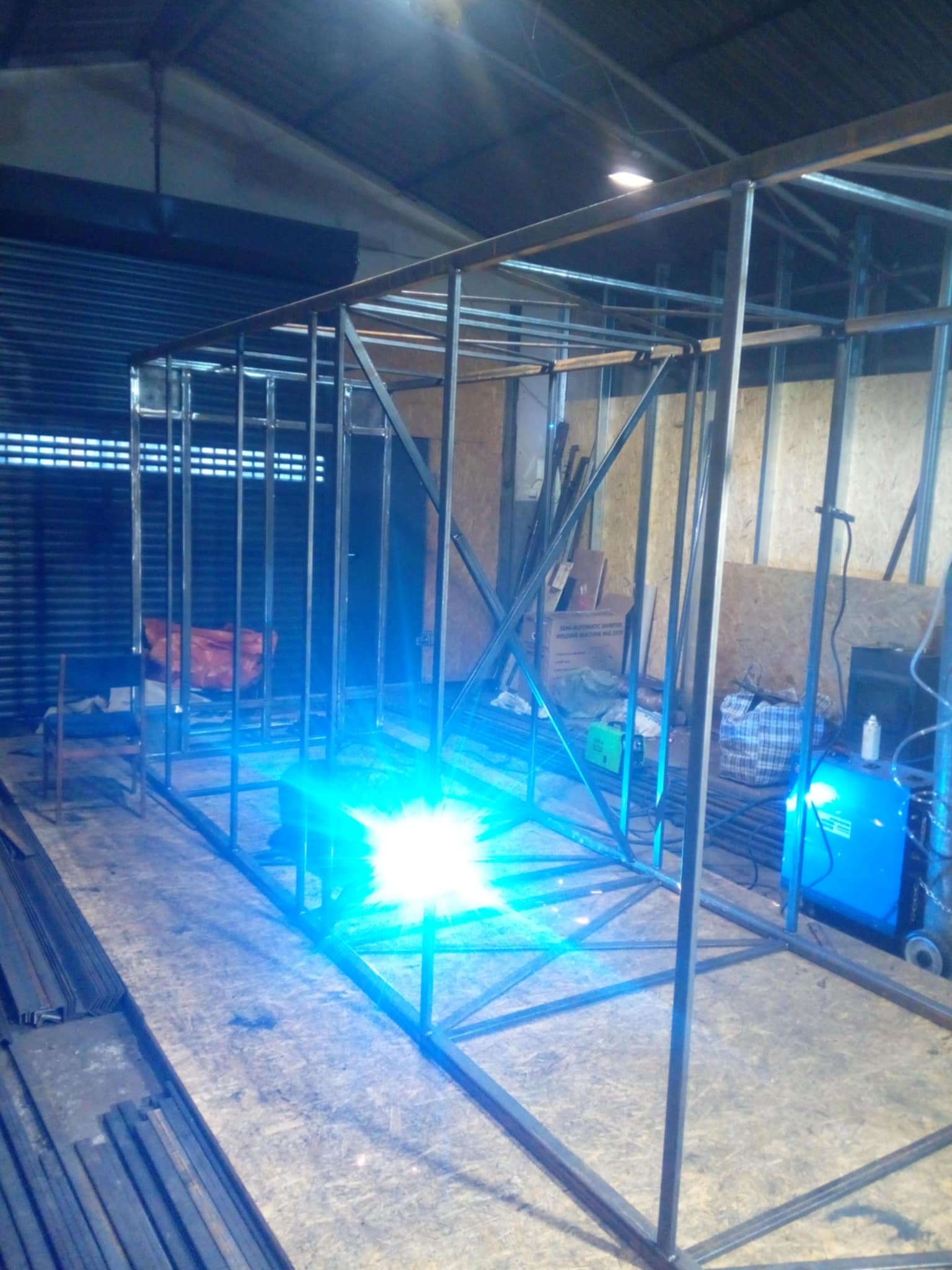 Konstrukcja stalowa w trakcie spawania, widoczny intensywny błysk spawarki, surowe otoczenie warsztatowe, elementy konstrukcyjne ułożone na płycie OSB.