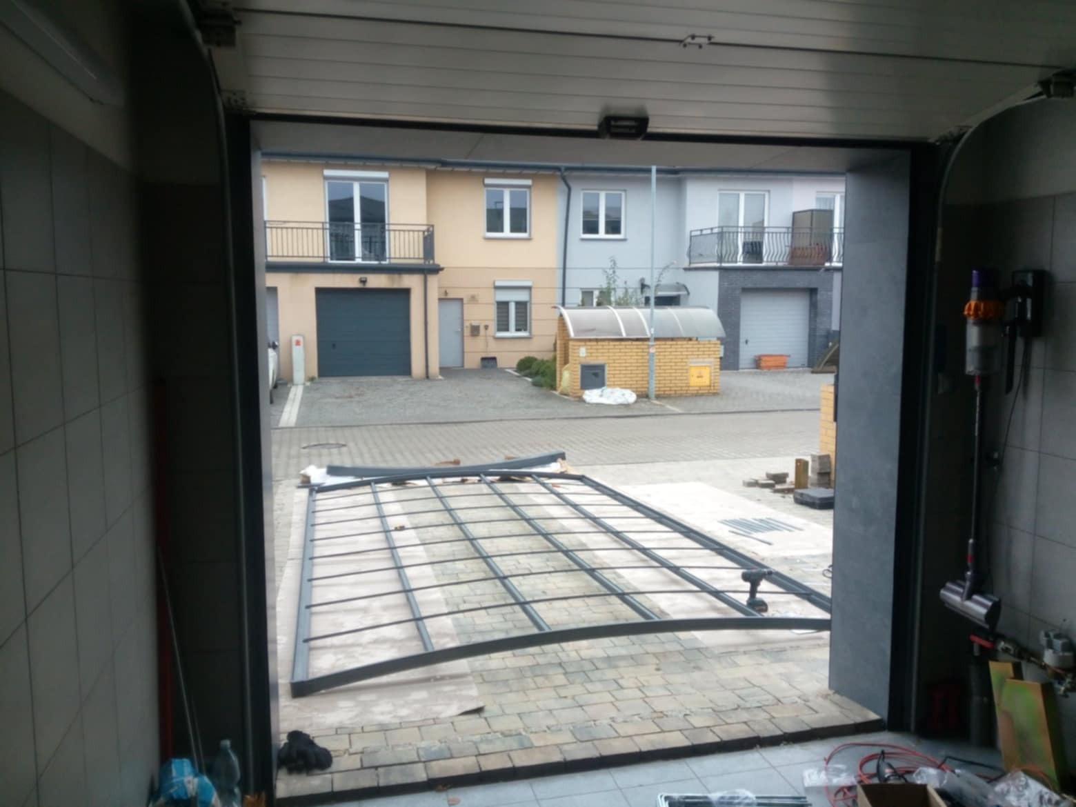 Wnętrze garażu z widokiem na ulicę, gdzie na podłodze leży częściowo zmontowana metalowa pergola, przygotowywana do montażu przed domem.