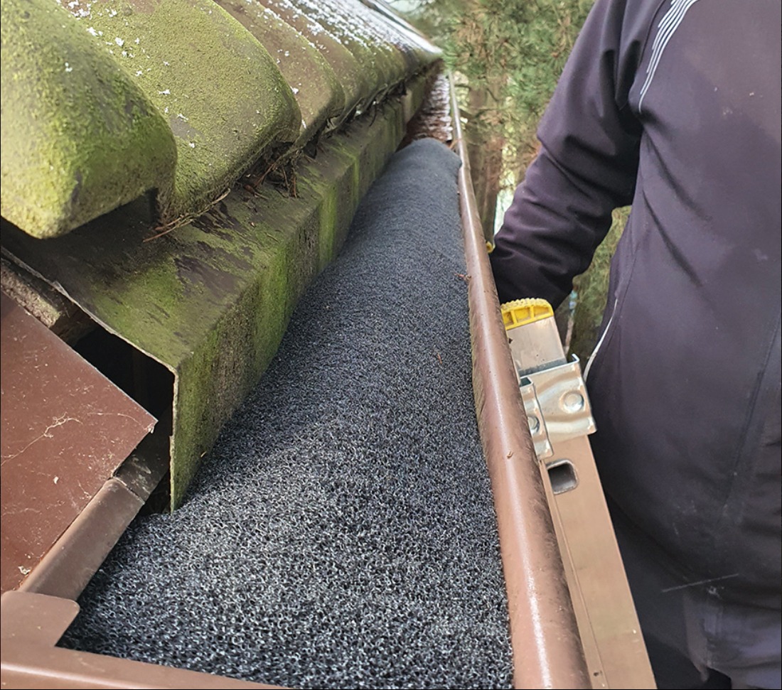 Rynna wypełniona szarym wkładem filtrującym z drabiny, widoczne mchy na dachu i fragment sylwetki osoby w ciemnej kurtce.