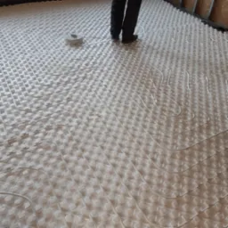 Biała mata izolacyjna z ułożonymi rurami ogrzewania podłogowego, widoczne nogi instalatora w czarnych spodniach i butach roboczych oraz fragment ściany w tle.