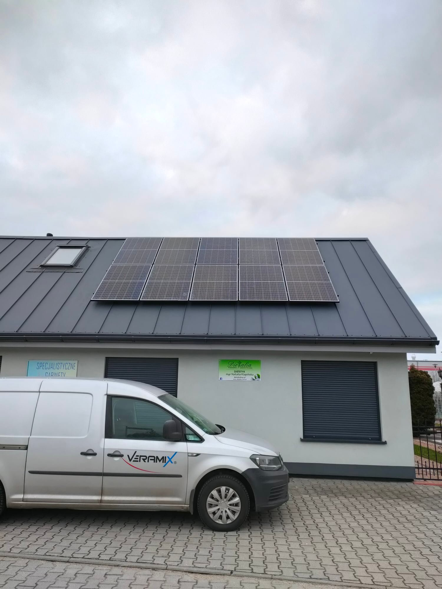 Panele słoneczne na szarym dachu budynku z blachy trapezowej, z boku zaparkowany biały samochód dostawczy z logo firmy.
