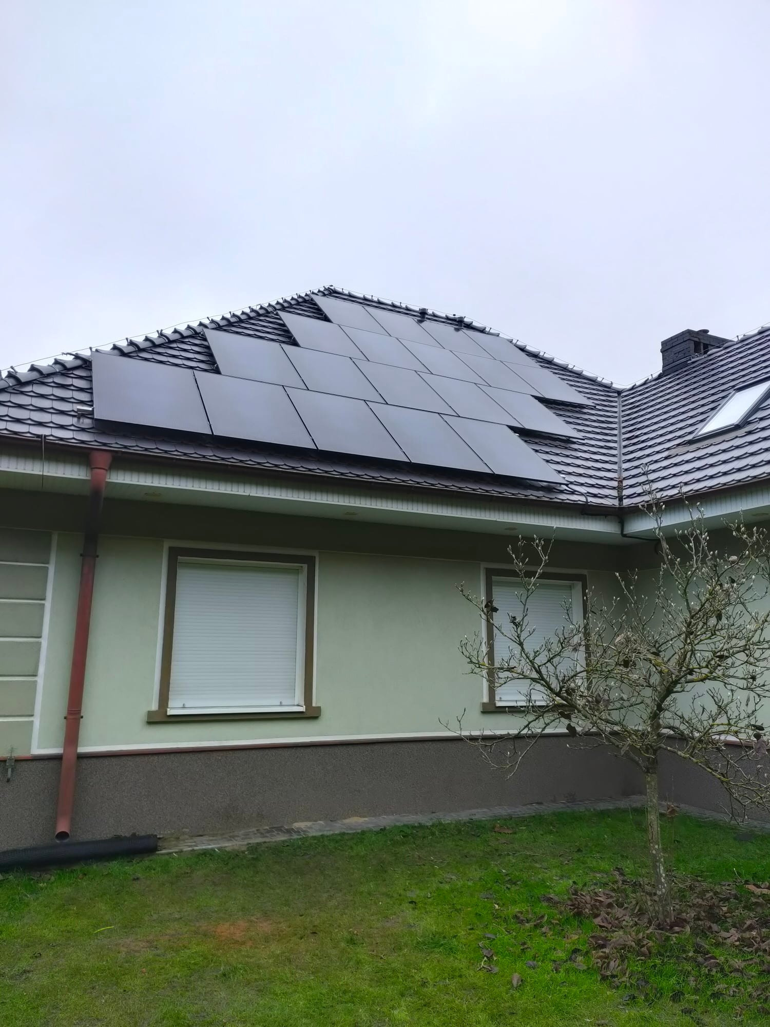 Panele fotowoltaiczne na dachu domu z brązową dachówką, widok na część elewacji z oknami z roletami zewnętrznymi i drzewem bez liści na trawniku.