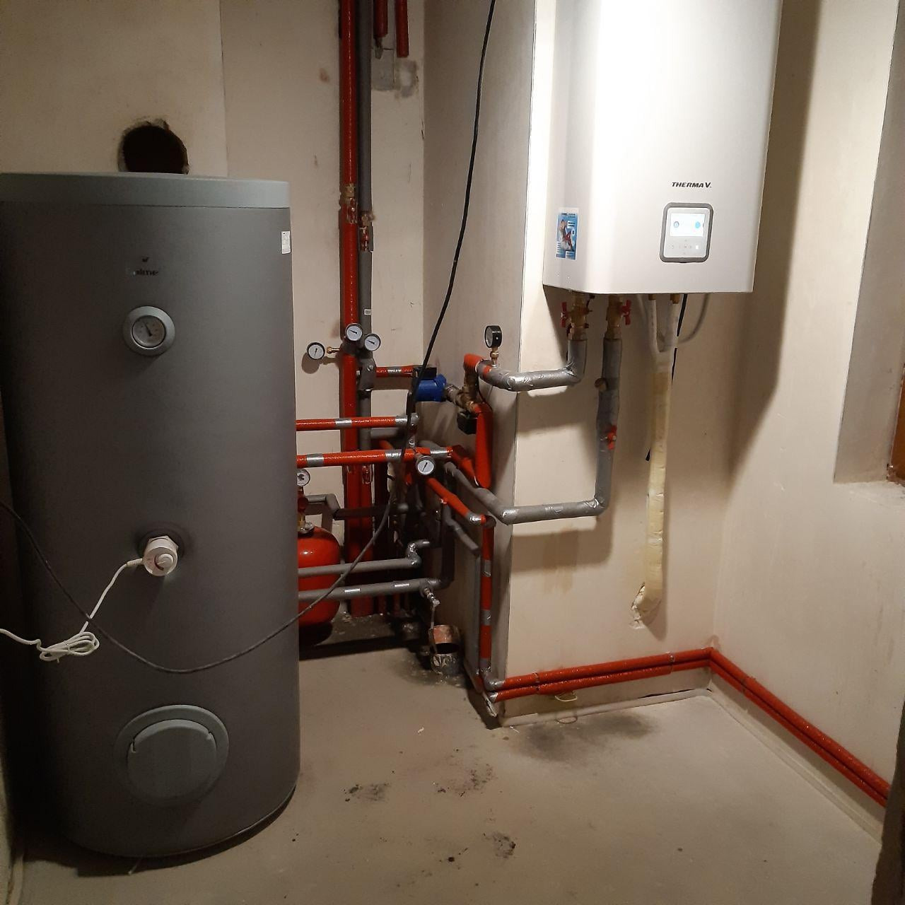 Zainstalowana biała jednostka wewnętrzna Therma V z szarym zbiornikiem akumulacyjnym w kotłowni z widocznymi rurami w izolacji koloru czerwonego.