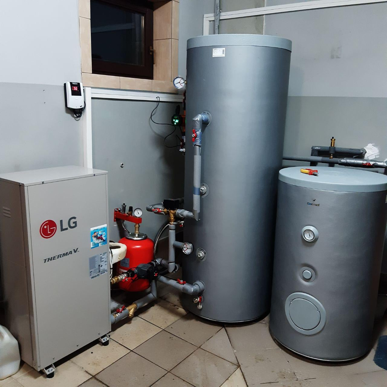 Instalacja grzewcza w kotłowni: zbiorniki akumulacyjne ciepła (jeden z narzędziem na górze) oraz jednostka zewnętrzna z logo LG Therma V, podłączone rurami z zaworami i manometrami.