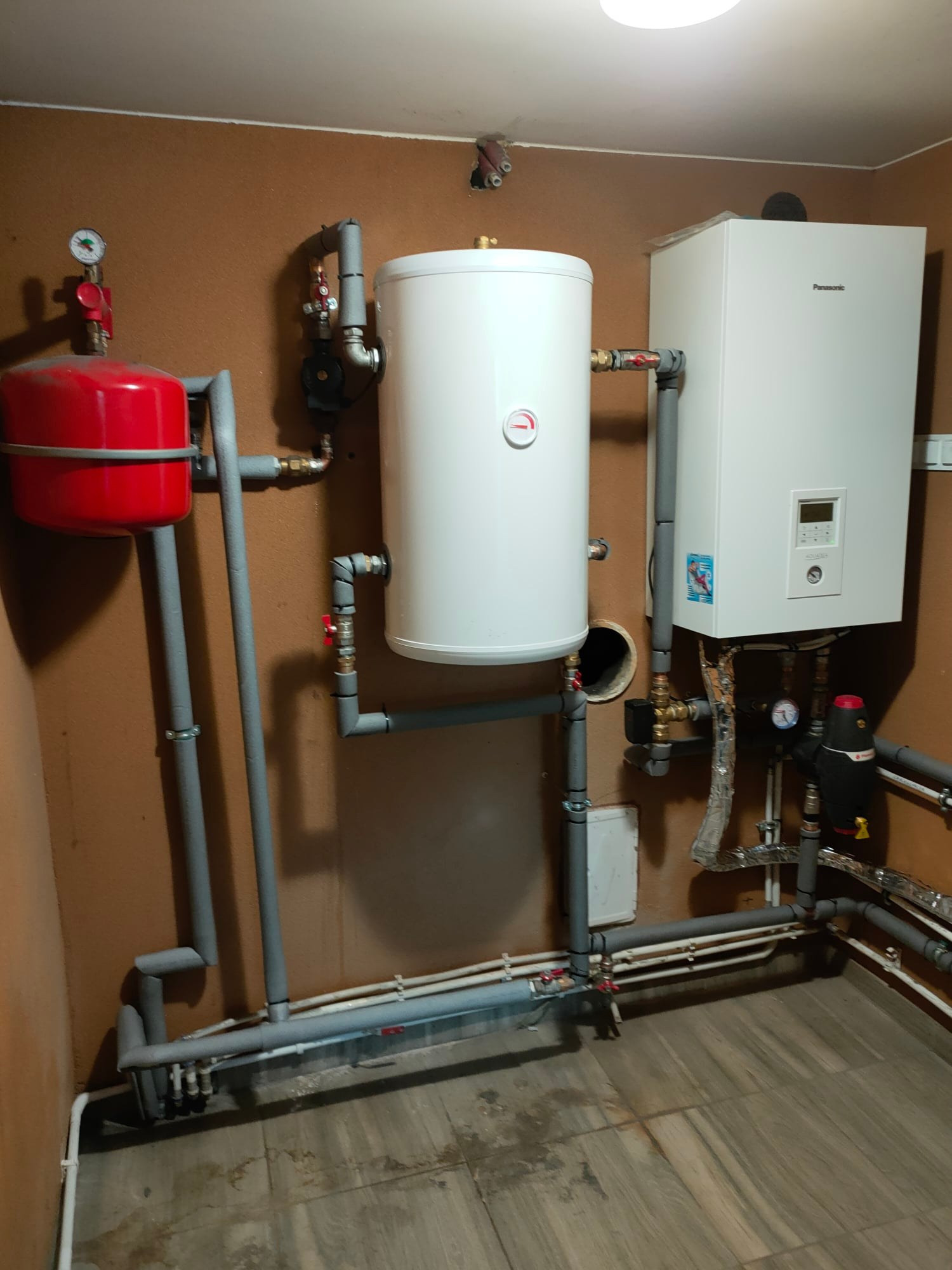Instalacja grzewcza z białym zasobnikiem ciepłej wody i wiszącym piecem grzewczym Panasonic, rury w izolacji, brązowe ściany, płytki podłogowe.